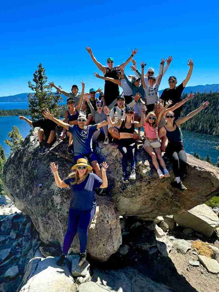 A large group of people posing together on a rocky outcrop with a scenic lake and forested mountains in the background.