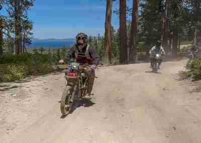 Two riders on electric dirt bikes on a dirt road through the pines, Lake Tahoe behind