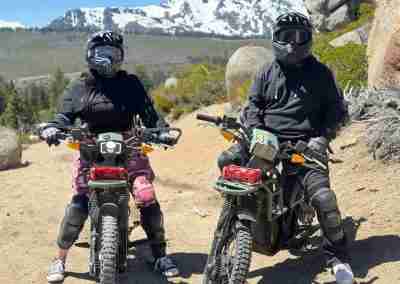 Two riders in helmets on electric dirt bikes with snowy peaks behind