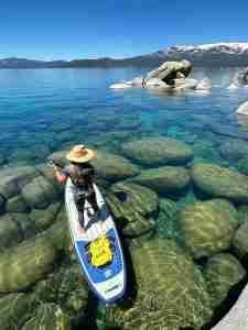 A serene scene of a paddleboarder in a sun hat gliding over the transparent blue-green waters of Lake Tahoe, with submerged boulders clearly visible below and distant snow-covered mountains against a clear sky.