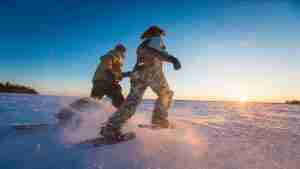 Two people snowshoeing across snowy field at sunset with snow spray in the air
